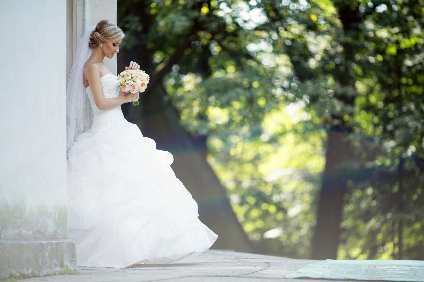 Beauty bride in bridal gown with bouquet and lace veil in the nature. Beautiful model girl in a white wedding dress. Female portrait in the park. Woman with hairstyle. Cute lady outdoors