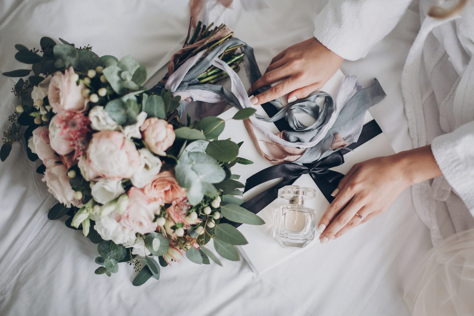 Modern wedding bouquet, perfume bottle, and gift box on white bed. Bride holding white box for morning boudoir before wedding ceremony. Wedding arrangements or bridal shower